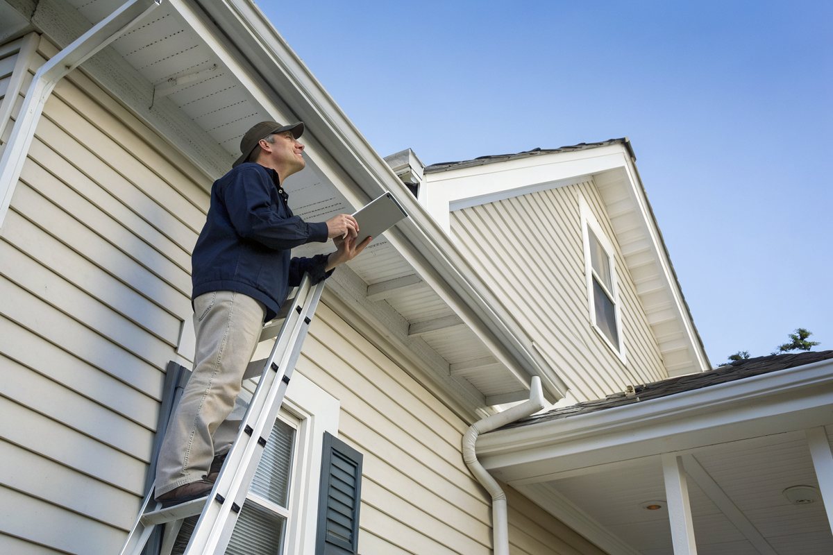 A man on a ladder checking the roof edge and gutter of a white-sided house
